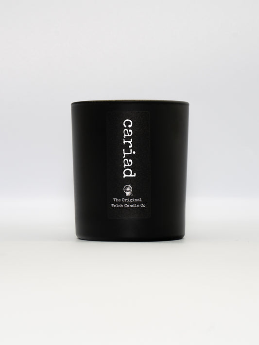 A matt black glass jar with a label saying cariad, filled with a black pomegranate soy candle
